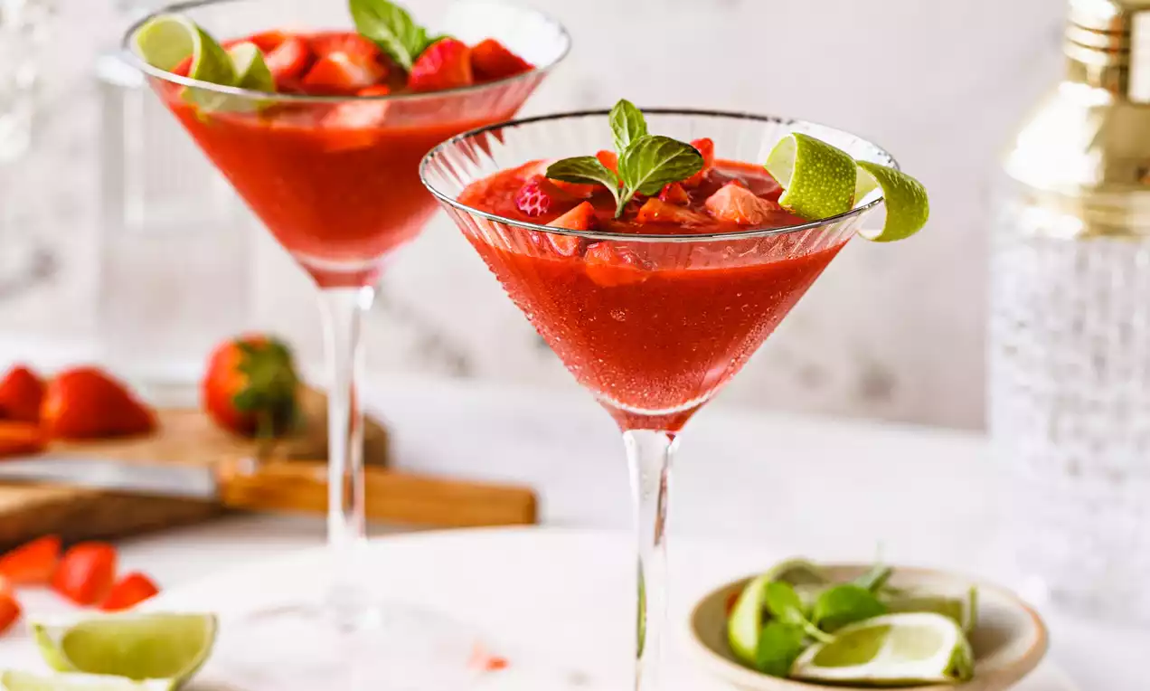 Picture - Two glass martini cups filled with vibrant red daiquiri, garnished with fresh strawberry slices, mint leaves and lime wedges on a white marble tray