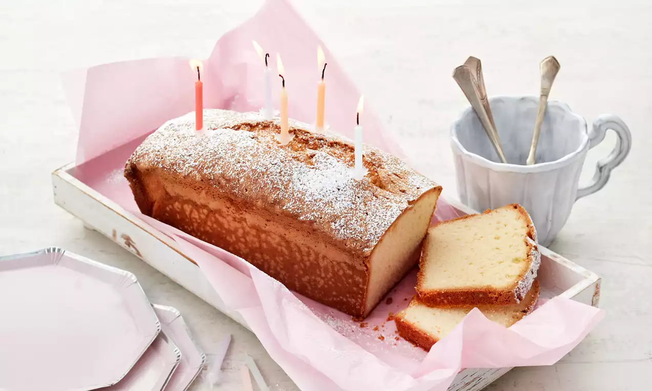Goldbrauner Sandkuchen mit feiner Puderzucker-Schicht, angeschnitten auf rosa Papier in weißer Holzschale, mit Kerzen dekoriert