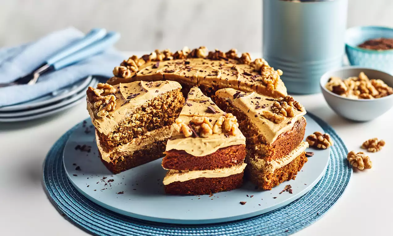 Layered coffee and walnut cake with light brown frosting, topped with walnut halves and chocolate shavings on a blue plate