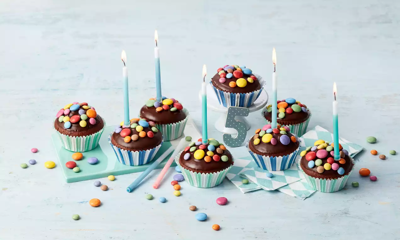 Schokoladenmuffins mit bunten Smarties in gestreiften Papierförmchen, teils mit blauen Kerzen und einer silbernen Zahl fünf dekoriert auf hellblauem Hintergrund