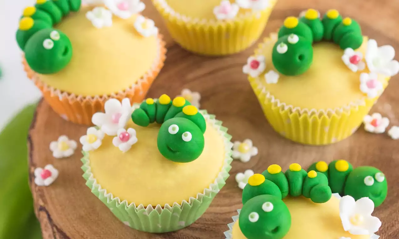 Babeczki w kolorowych papilotkach z gładkim żółtym kremem, ozdobione zielonymi gąsienicami z lukru i cukrowymi kwiatkami