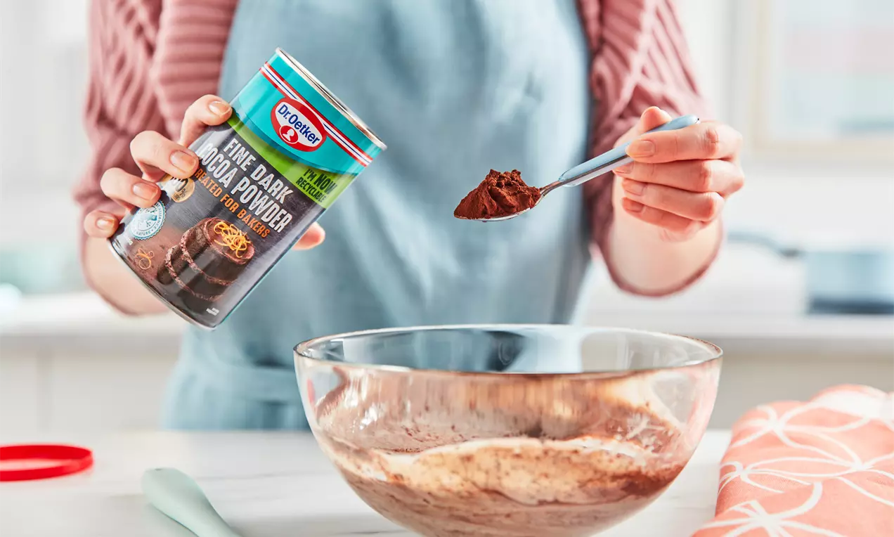 A glass bowl filled with light chocolate custard, a spoonful of deep brown cocoa powder being added from a tin by hands in a blue apron