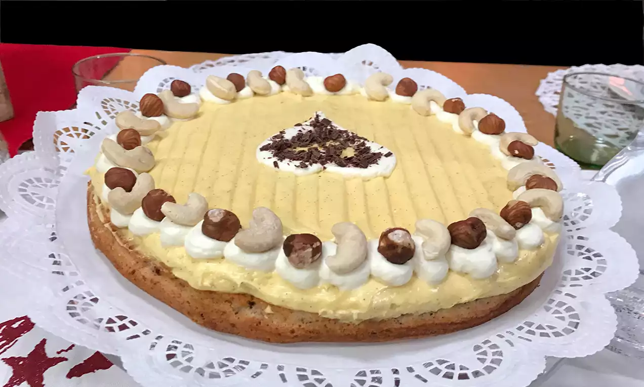 Goldbrauner Mandelkuchen mit gelber Creme, dekoriert mit Sahnetupfen, Haselnüssen und Cashewkernen auf weißer Spitzendecke