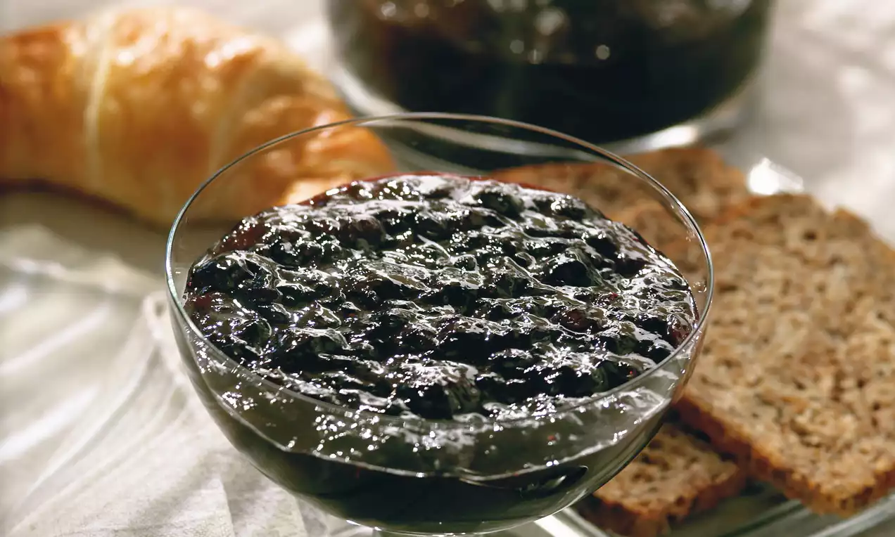 Confiture noire brillante aux reflets profonds dans un bol en verre avec des tranches de pain brun et un croissant doré en arrière-plan