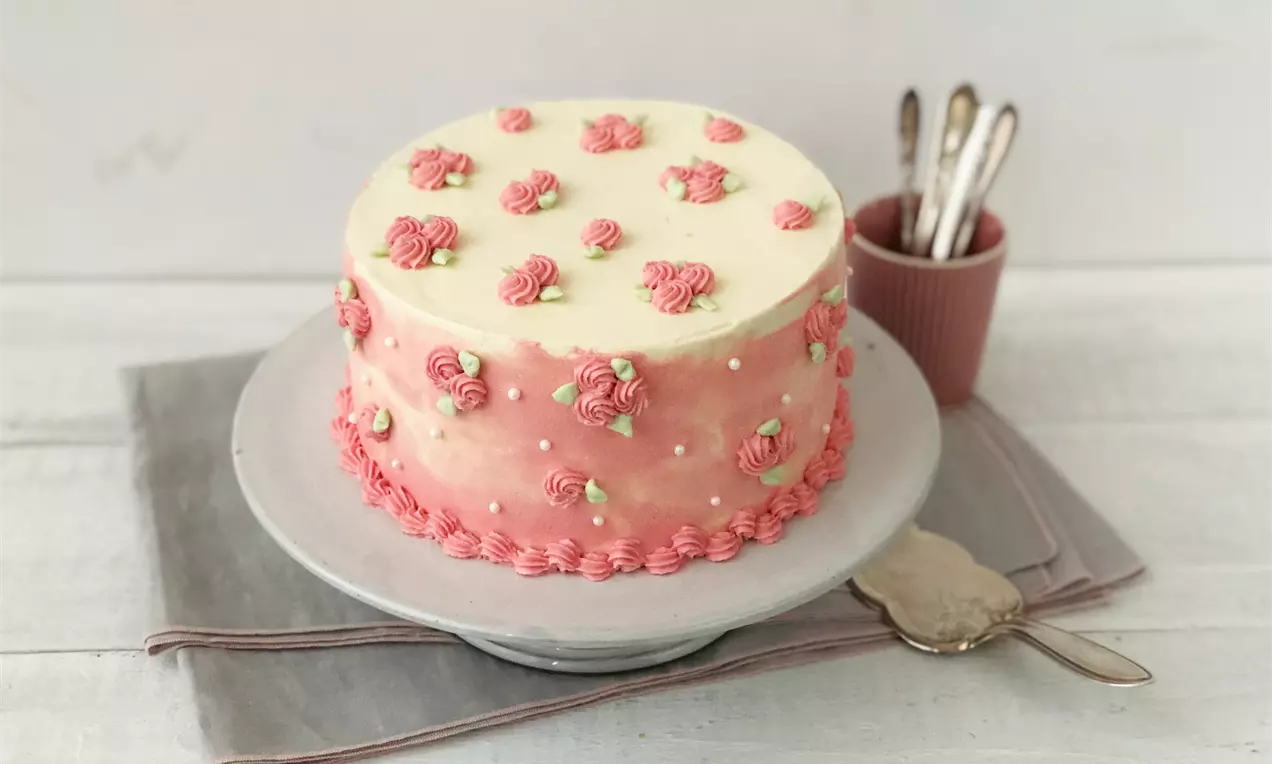 Zweischichtige Torte mit rosa Farbverlauf, dekoriert mit kleinen rosa Rosen und grünen Blättern, auf einer weißen Platte mit Besteck im Hintergrund