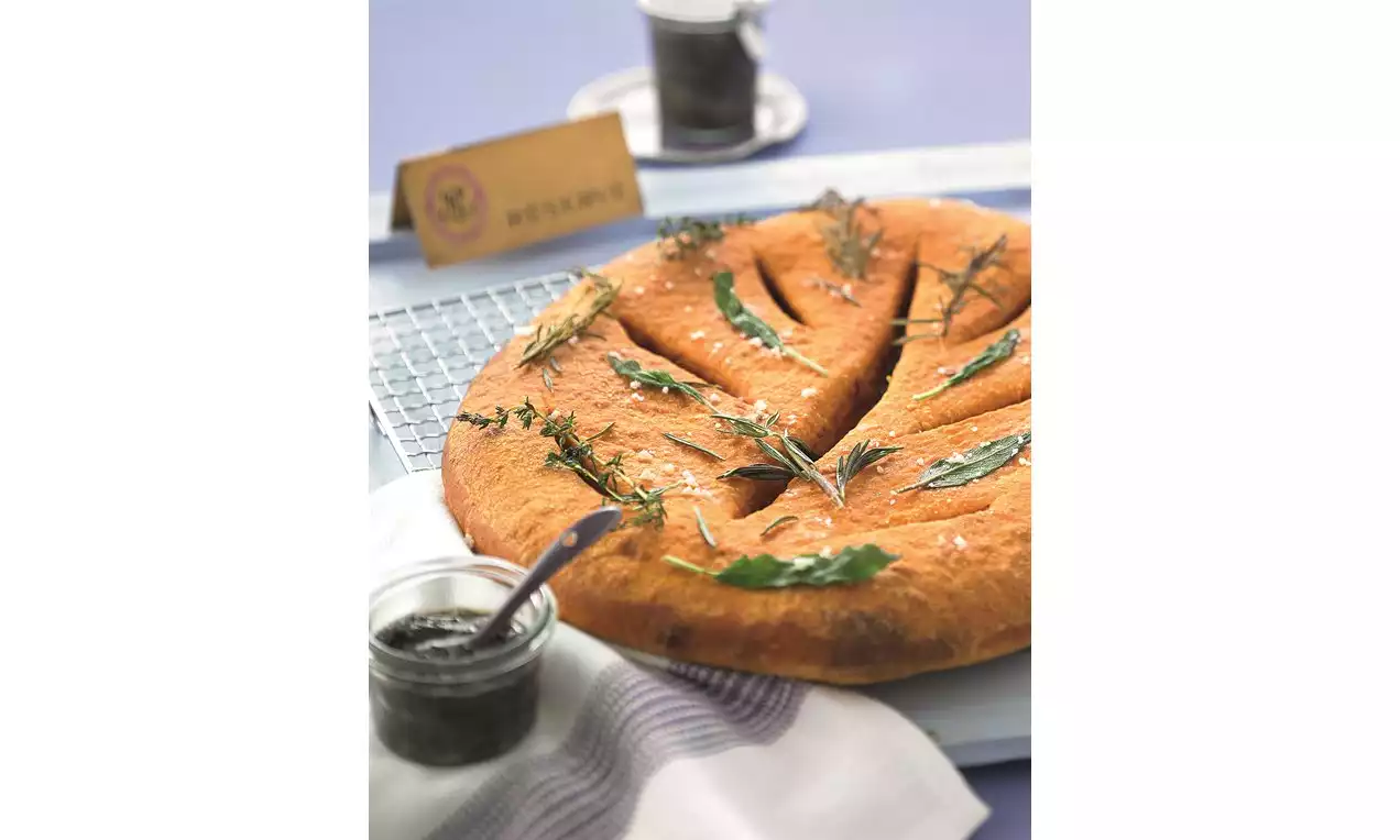 Fougasse z zlato rjavo skorjo v obliki listnatih izrezov, okrašena s svežimi zelišči, postrežena ob steklenem lončku tapenade na beli krpi