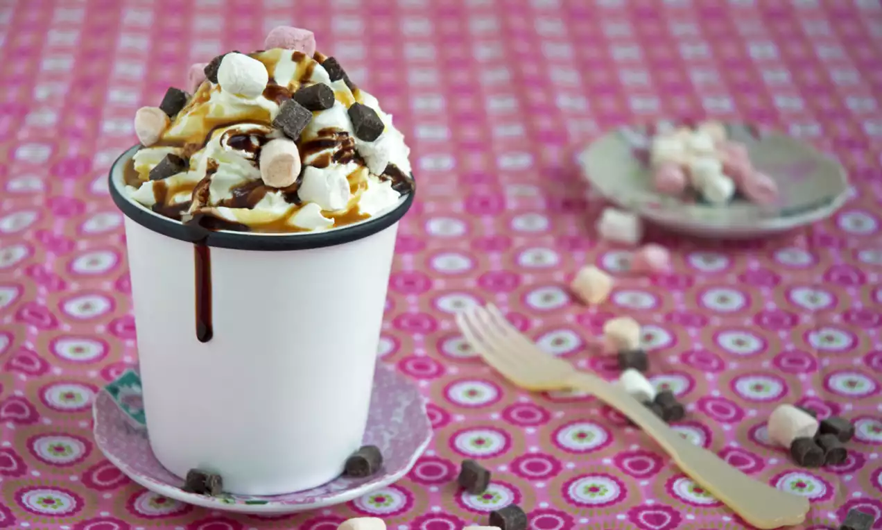 Witte mok gevuld met luchtige mugcake, afgetopt met slagroom, kleine marshmallows, chocoladestukjes en karamelsaus, op een kleurrijk tafelkleed