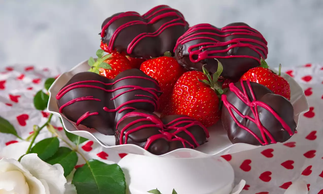 Srdcové jahody namočené v tmavé čokoládě, s růžovými čokoládovými pruhy, uspořádané na bílém podnosu vedle čerstvých červených jahod