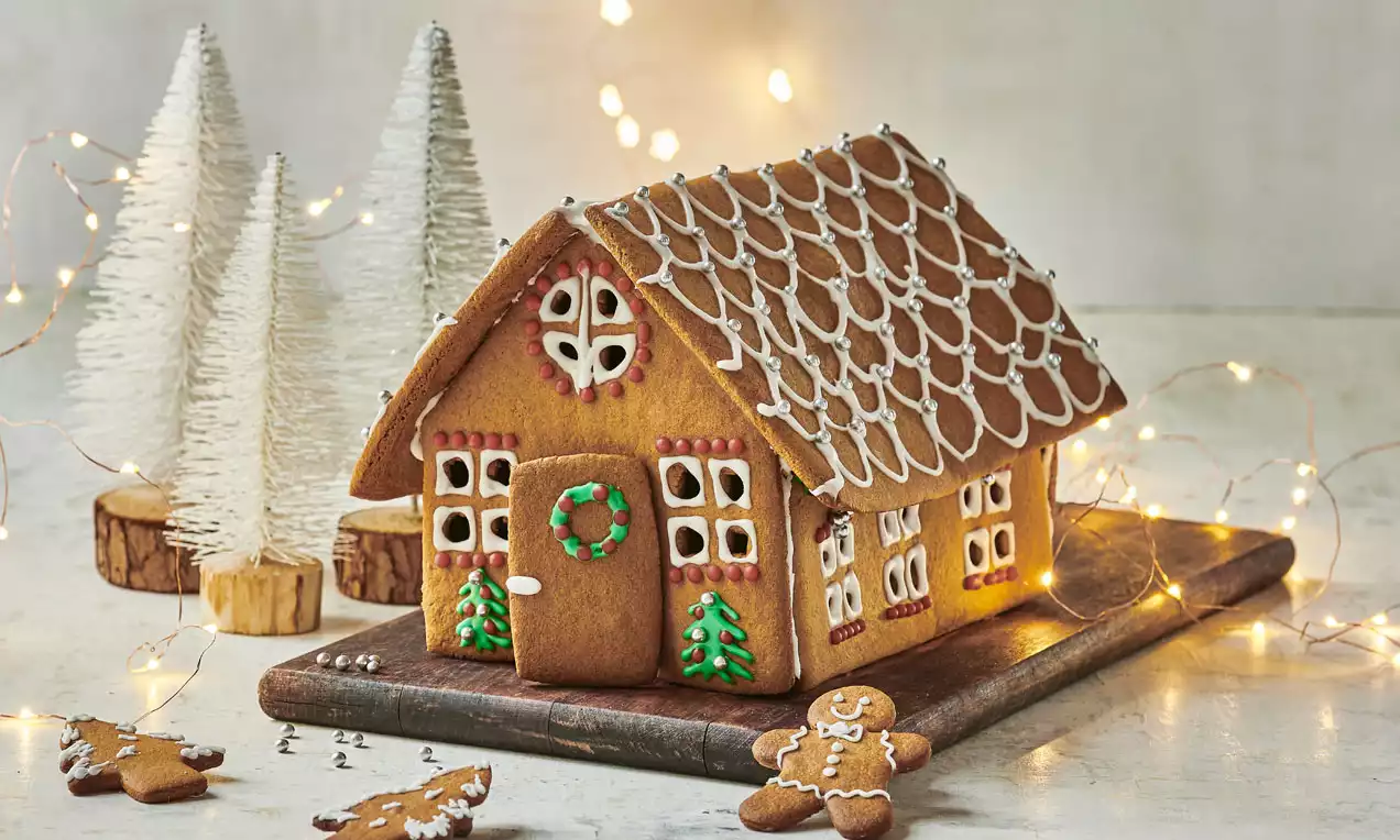 Ein Honigkuchenhaus mit weißer Zuckerglasur, bunten Details und Verzierungen, umgeben von Lebkuchenfiguren auf einem Holzbrett