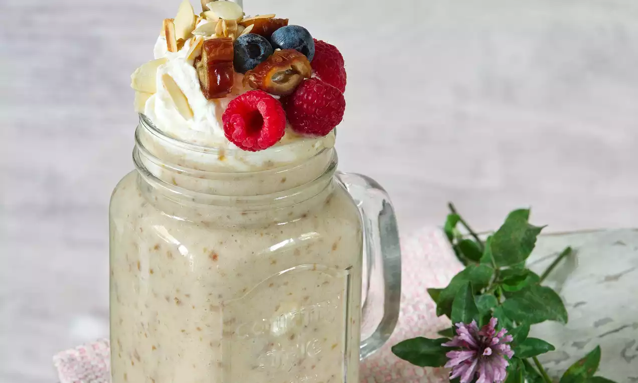 Banánový koktejl světle béžové barvy v průhledné sklenici s bílou šlehačkou, malinami, borůvkami, mandlemi a květinovou dekorací vedle