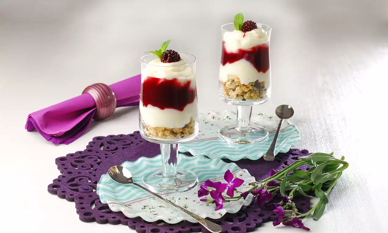 Cam kup bardaklarında sunulan, altı çıtır bisküvi tabanlı, kremsi beyaz katmanlı ve parlak böğürtlen sosuyla süslenmiş tatlı