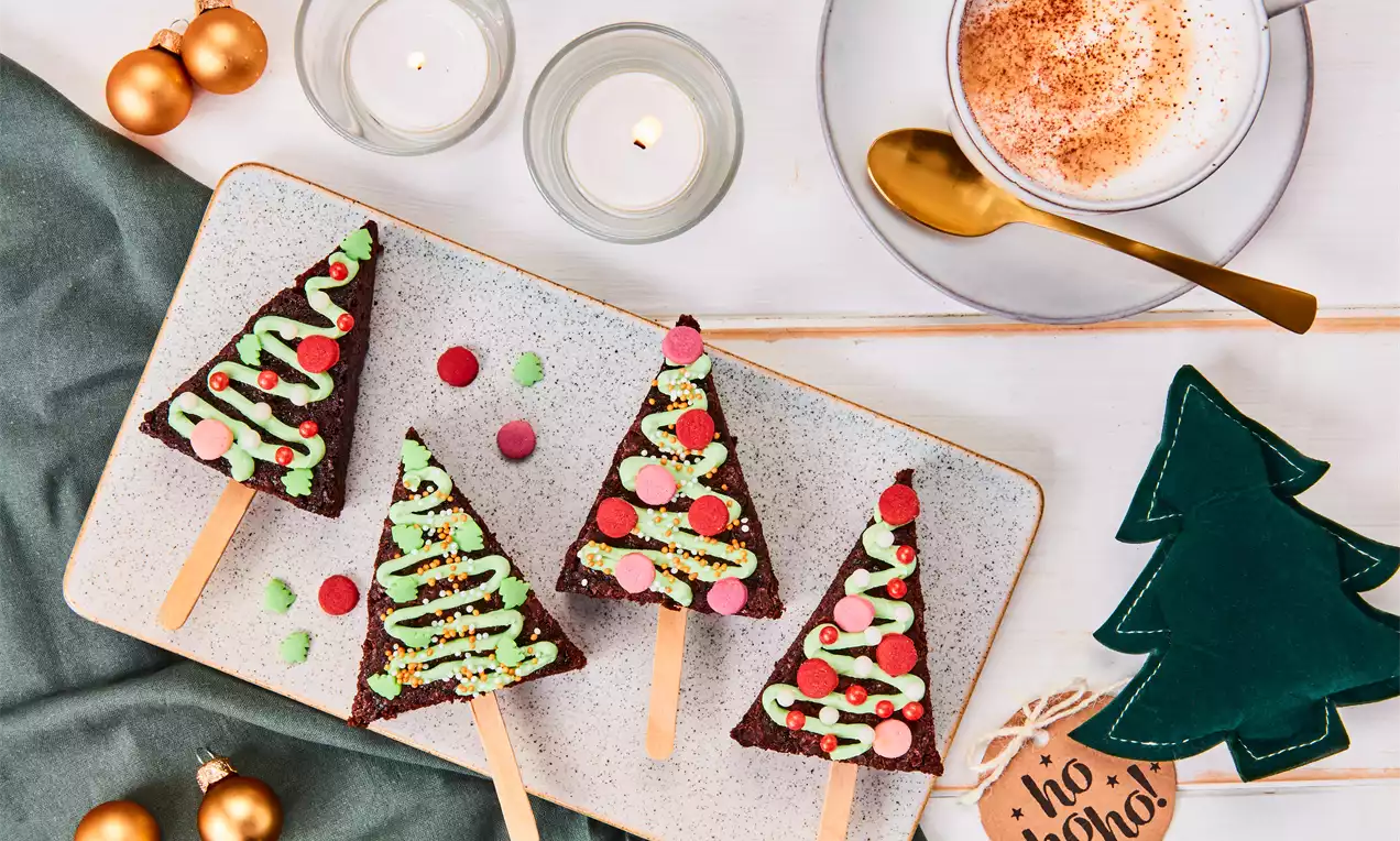 Brownies en forme de sapins avec glaçage vert, décorés de perles et confettis colorés sur bâtonnets en bois sur une assiette blanche