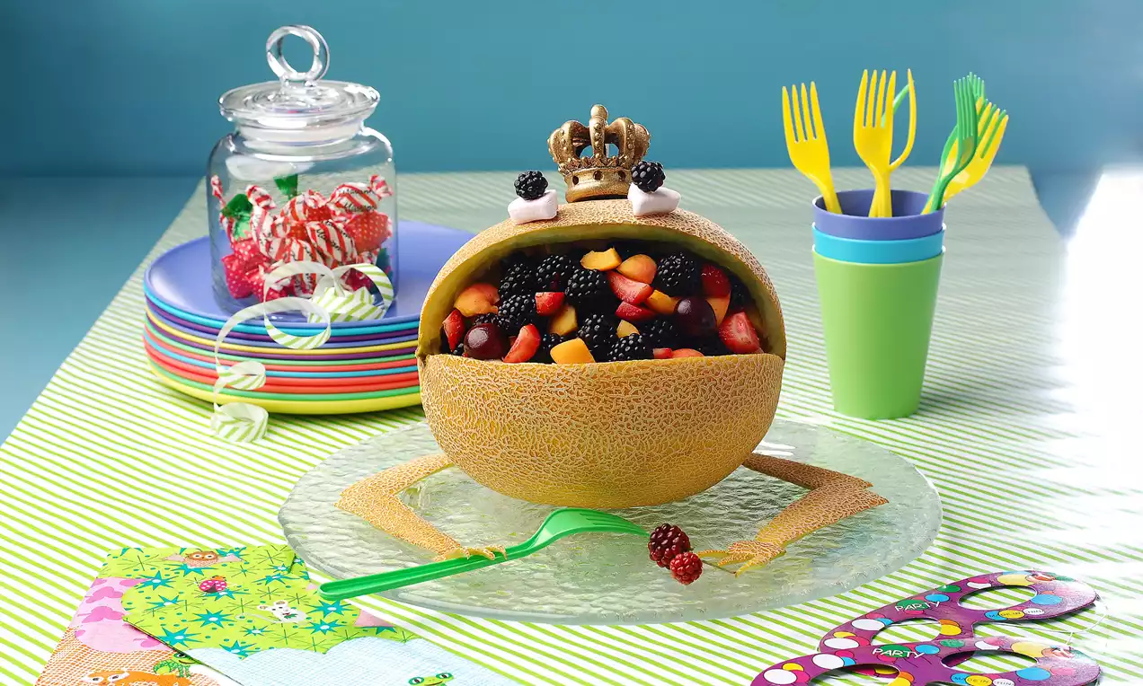 Kavundan yapılmış kurbağa şekli, içi renkli meyvelerle dolu, altın taçlı, cam tabak üzerinde renkli masa düzeniyle sunulmuş