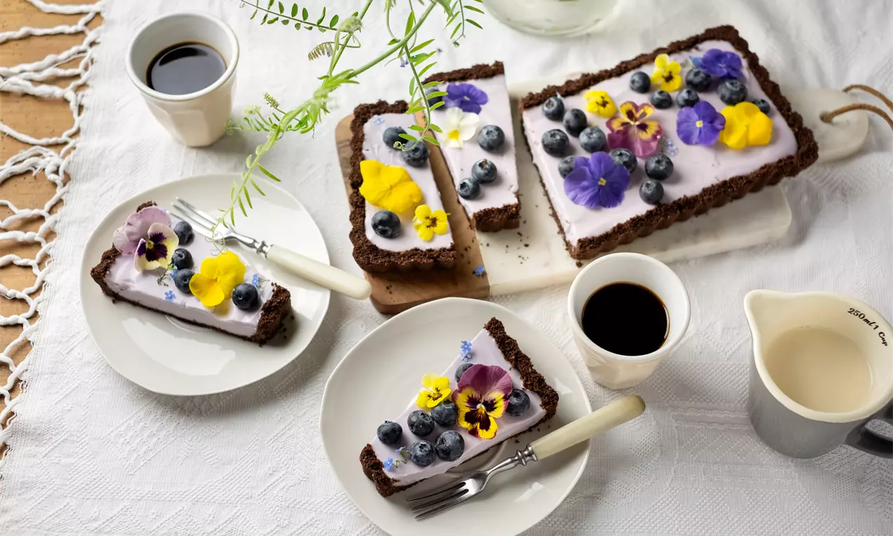 Тарта с правоъгълна форма, шоколадов блат, бледолилав крем, пресни боровинки и цветчета виола върху плато, сервирана на маса