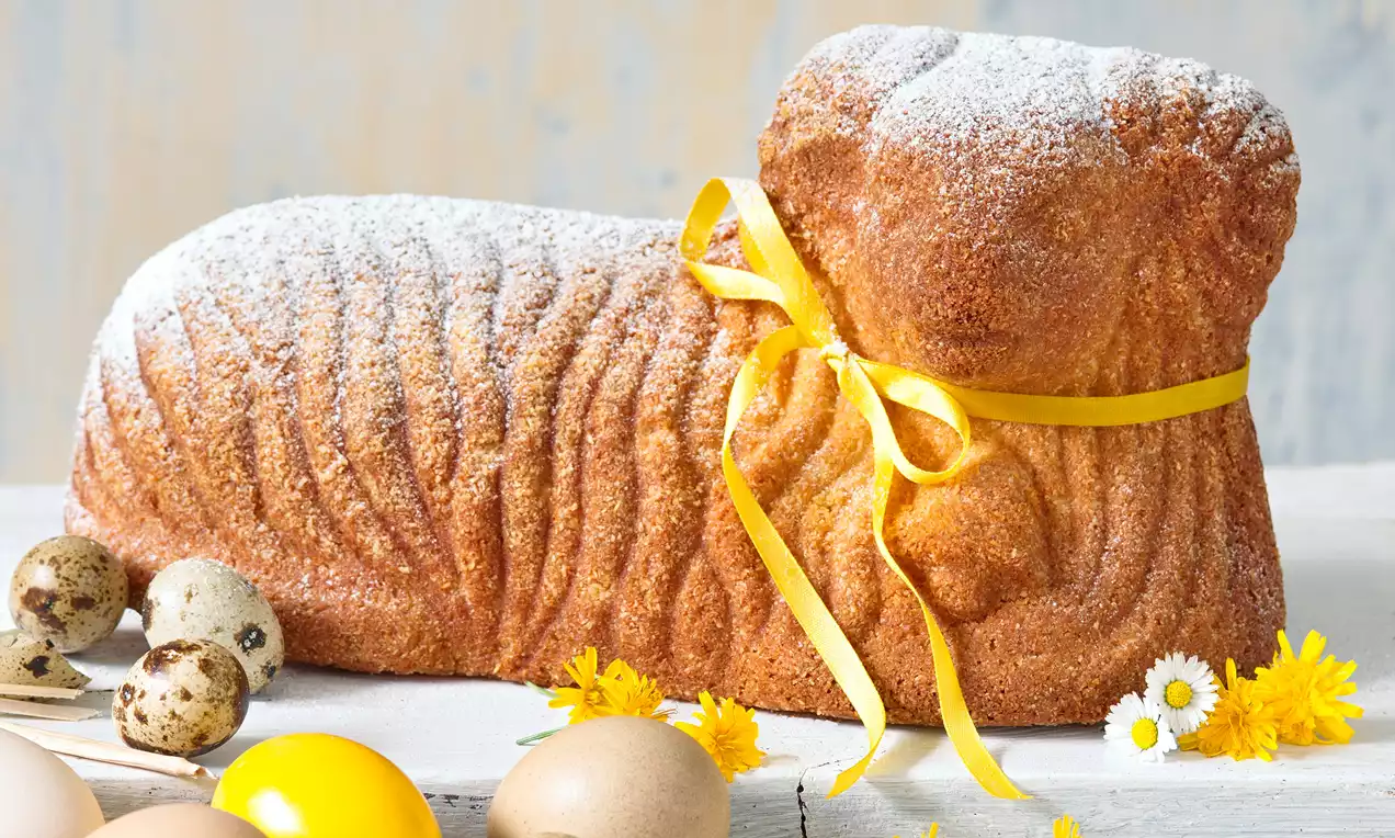 Světle hnědý třený beránek s jemnou texturou, posypaný moučkovým cukrem, ovázaný žlutou stuhou, na bílém dřevěném stole obklopený vajíčky a květinami