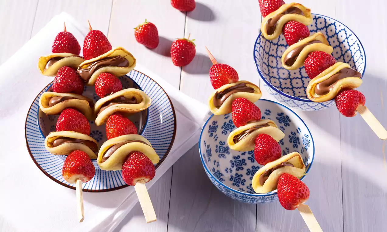 Çıtır pembe çilekler ve katlı yumuşak krepli pancake dilimleri ahşap çubuklarda sırayla dizilmiş, seramik kaselerde sunulmuş