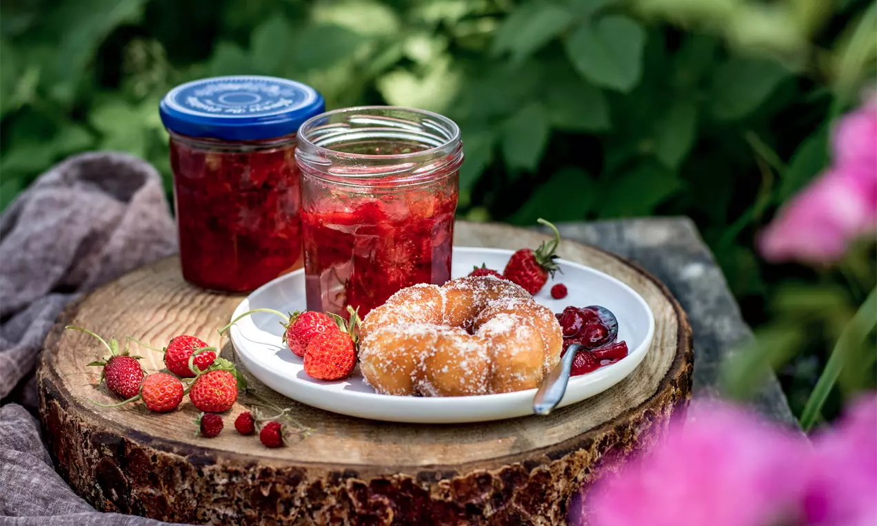 Яркочервено ягодово сладко с фина текстура в стъклен буркан до чиния с поръсени с пудра захар кифлички и свежи ягоди върху дървена поставка