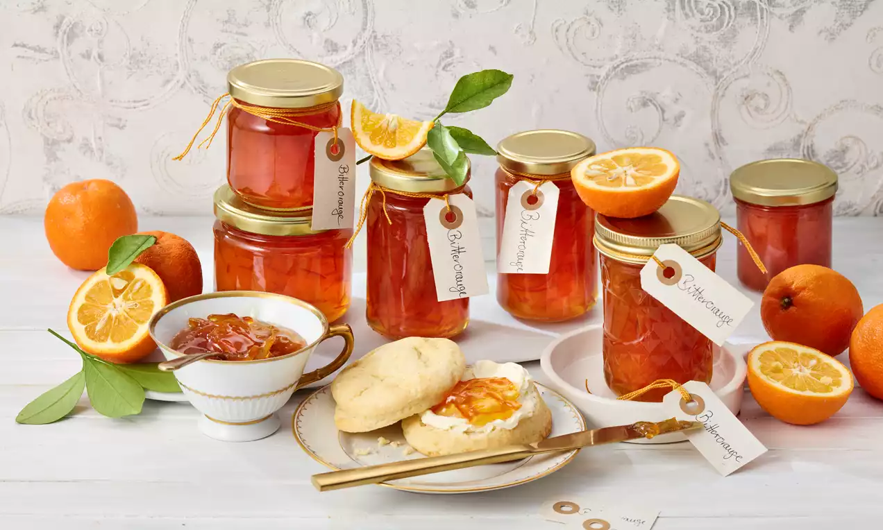 Goldorange Bitterorangenmarmelade in klaren Gläsern mit Deckeln und Etiketten, umrahmt von frischen Orangen, Gebäck und Marmeladenschale