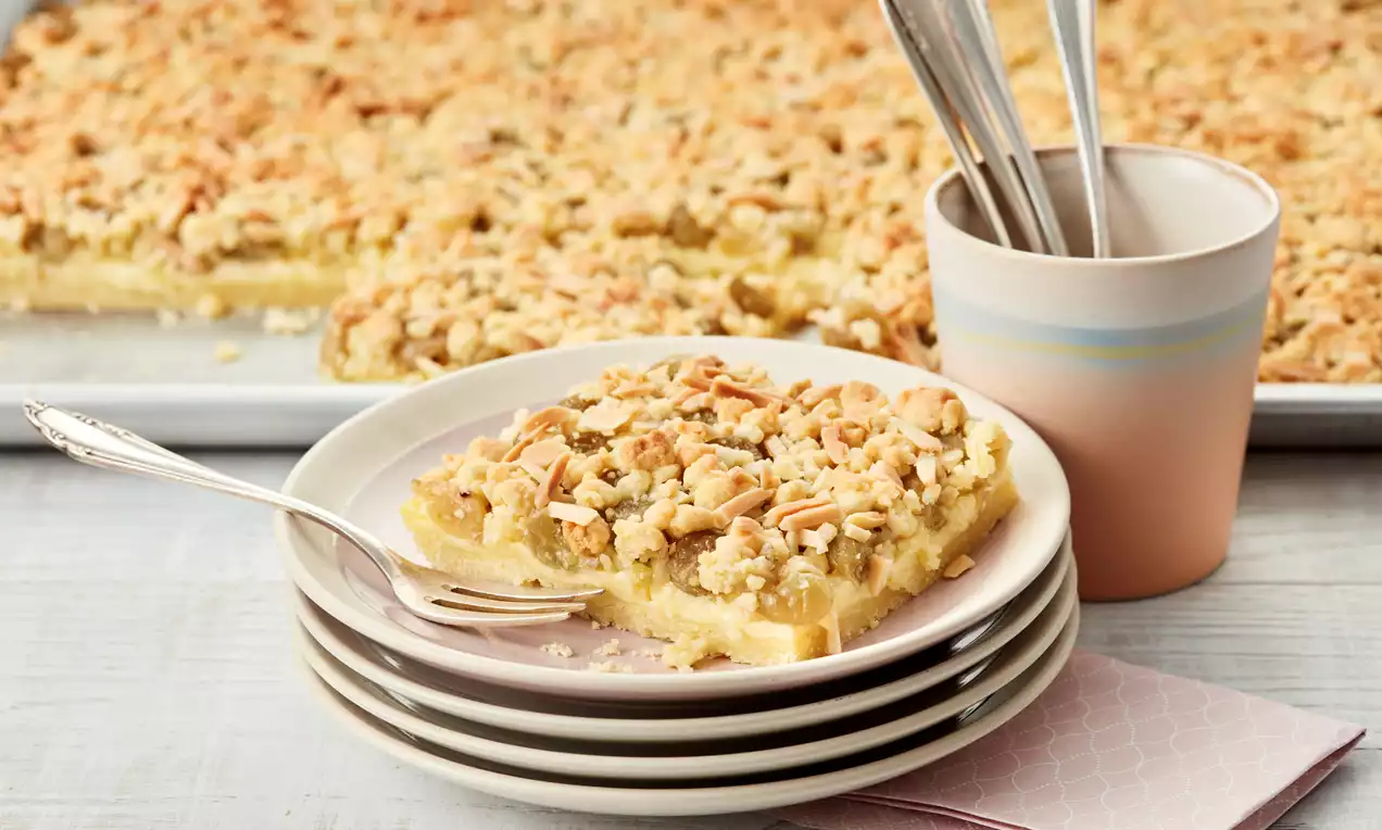 Stachelbeerkuchen mit hellgelber Puddingfüllung, goldbraunen Streuseln und gehackten Mandeln auf einem weißen Teller mit Besteck und rosa Serviette