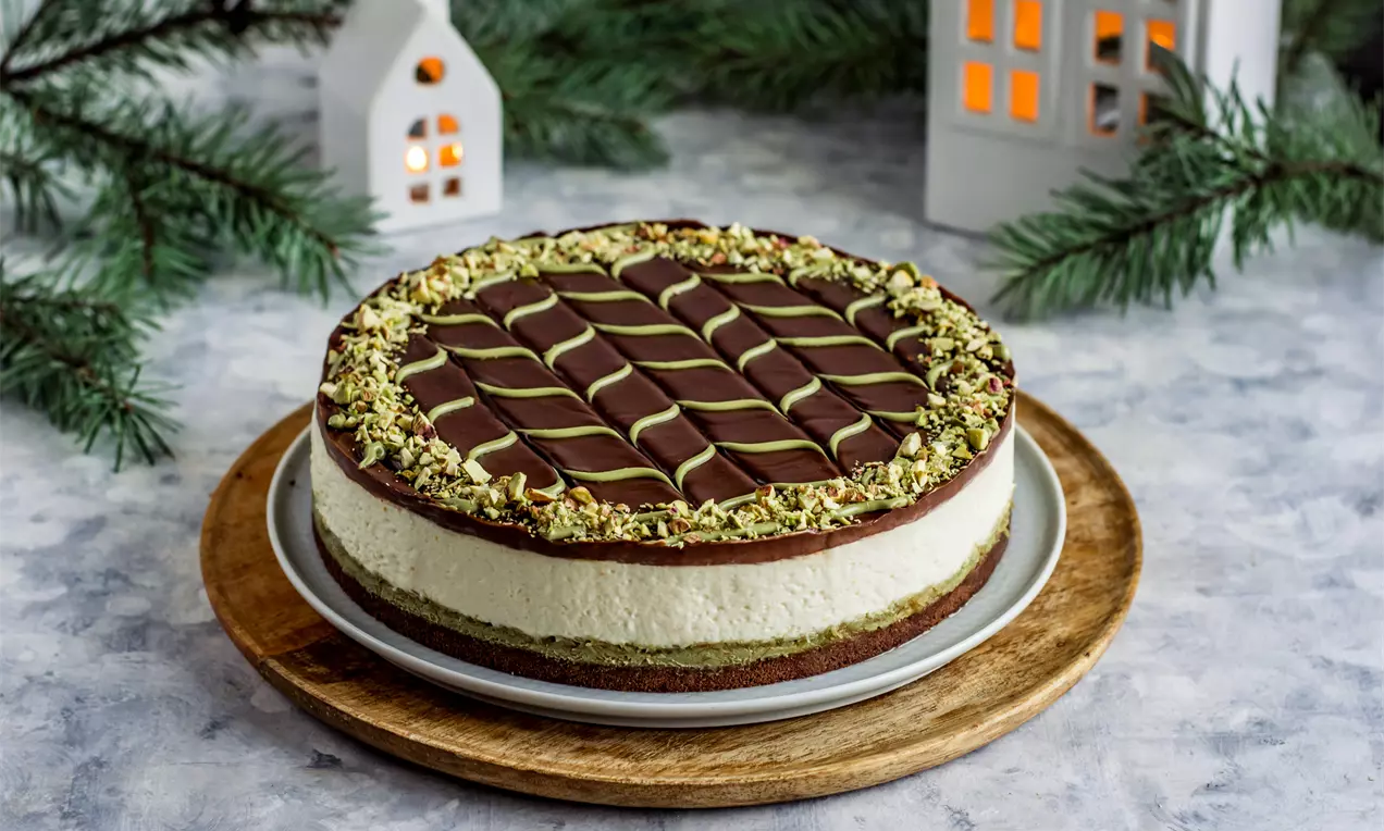 Чийзкейк с кремообразен бял пълнеж, кафяв блат, глазура с шоколадова решетка и зелен куполовиден кант от натрошени шамфъстъци върху дървена поставка