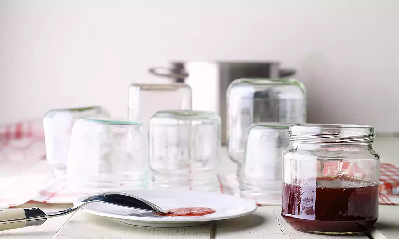 Roter Fruchtaufstrich auf einem weißen Teller neben einem Glas mit rubinrotem Gelee, umgeben von umgedrehten Gläsern auf heller Holzfläche
