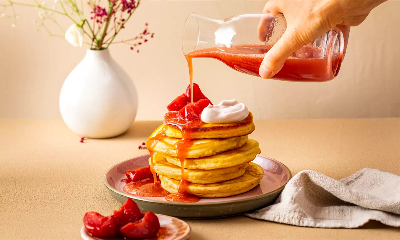 Жълти пухкави палачинки на купчина, полети с червено сливово пюре, гарнирани с крем с резени сливи върху розова чиния