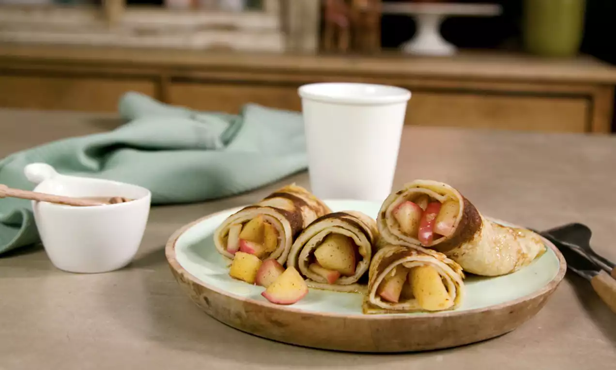 Pancakes roulés dorés garnis de morceaux de pommes cuites aux teintes dorées et rosées, disposés sur une assiette beige avec fond vert