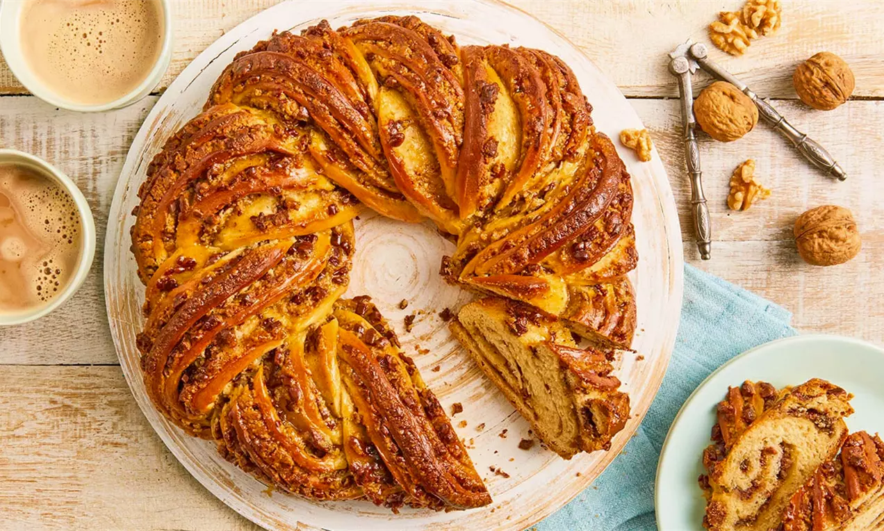 Кръгъл козунак кехлибаренокафяв с орехови парченца, изплетен спираловидно, поднесен върху бяла чиния до чаши кафе и натрошени орехи