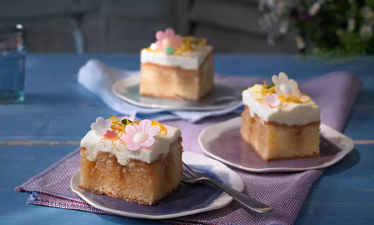 Saftige Kuchenwürfel mit hellem Teig, weißer Cremehaube, kandierten Blüten und gelben Zesten auf violetten Tellern arrangiert