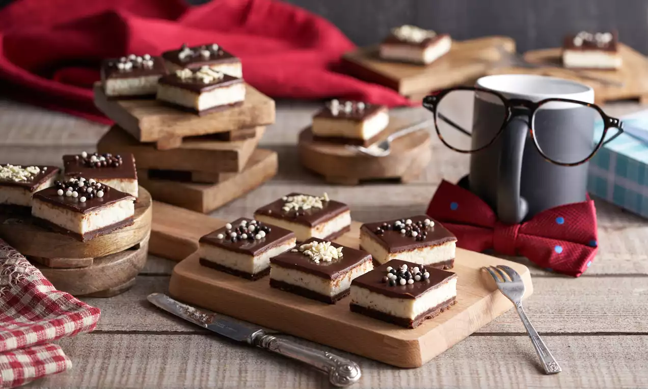 Tranci di cheesecake rettangolari con strati di crema chiara e cioccolato scuro, decorati con praline e cioccolato bianco su vassoi di legno
