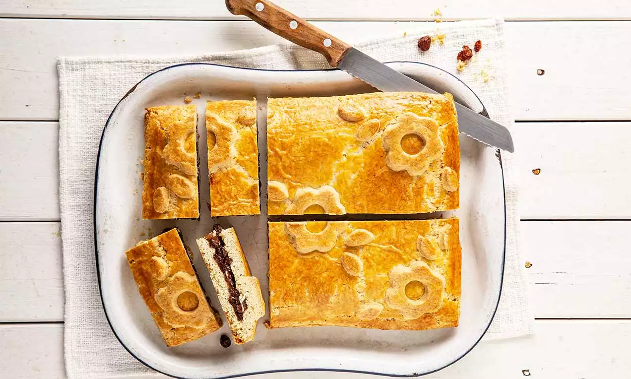 Goldgelbe rechteckige Urner Pastete mit dekorativen Blumenmustern, gefüllt mit dunkler Fleischfarce und heller Masse, auf weißer Platte mit Messer