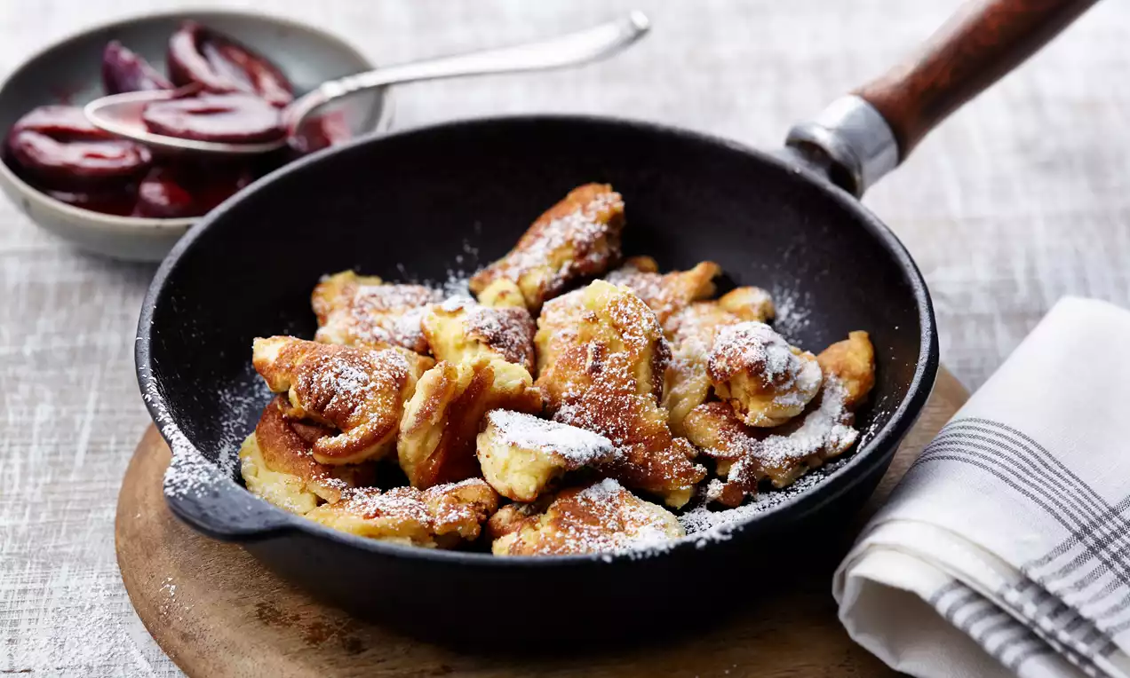Goldbraune, fluffige Kaiserschmarrnstücke mit weißem Puderzucker bestäubt in einer schwarzen Pfanne auf einem Holzbrett mit Zwetschgenkompott in Schale