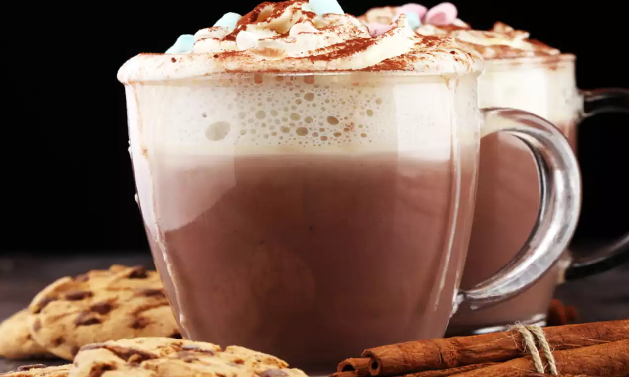 Hot chocolate in a clear glass mug with frothy cream, cocoa dusting, pastel mini marshmallows, cookies and cinnamon sticks beside it