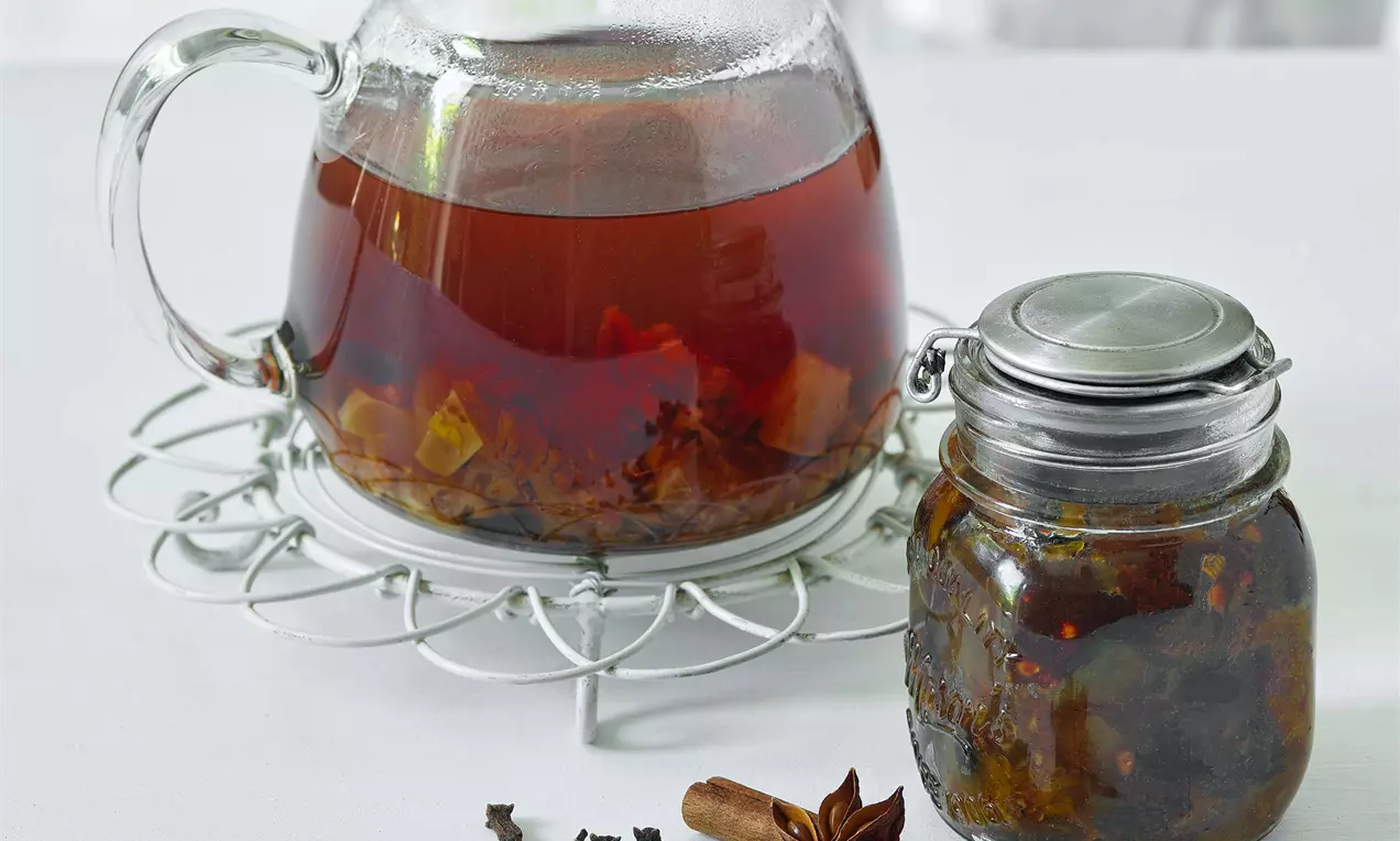 Tmavě jantarový šípkový čaj v průhledné skleněné konvici na kovovém podstavci vedle malé sklenice s víčkem, zdobené badyánem a skořicí