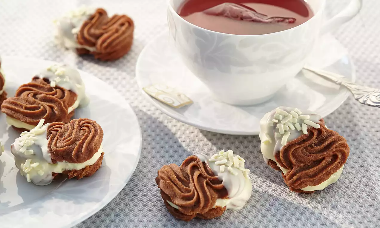 Petits biscuits torsadés couleur chocolat garnis de crème claire, trempés en partie dans du glaçage blanc et parsemés de pépites sur une assiette blanche
