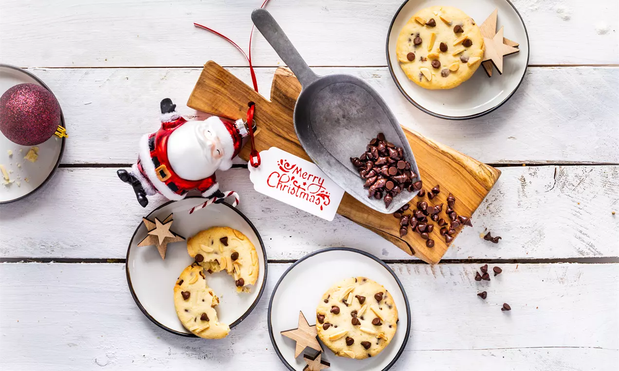 Кръгли маслени сладки с шоколадови капки, подредени в бели чинии с дървени звездички върху светъл фон до метална лопатка