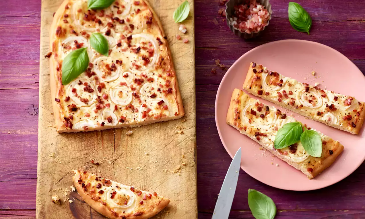 Knuspriger, rechteckiger Flammkuchen mit hellbraunem Teig, saftigen Zwiebelringen, Speckwürfeln und frischen Basilikumblättern auf Holzbrett