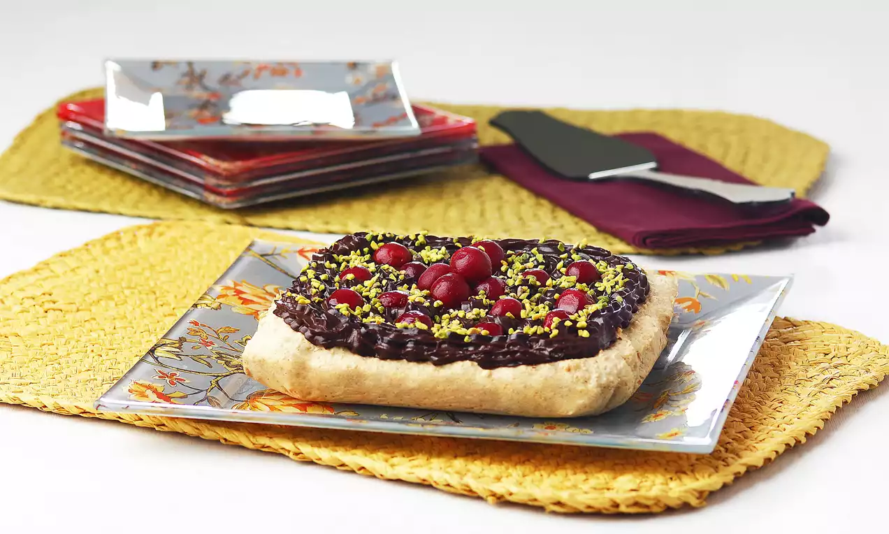 Kare desenli tabakta, kremalı fıstık ve kiraz ile süslenmiş kuyruğu yukarı kalkık, hafif kabarık hamur tabanlı çikolatalı tart