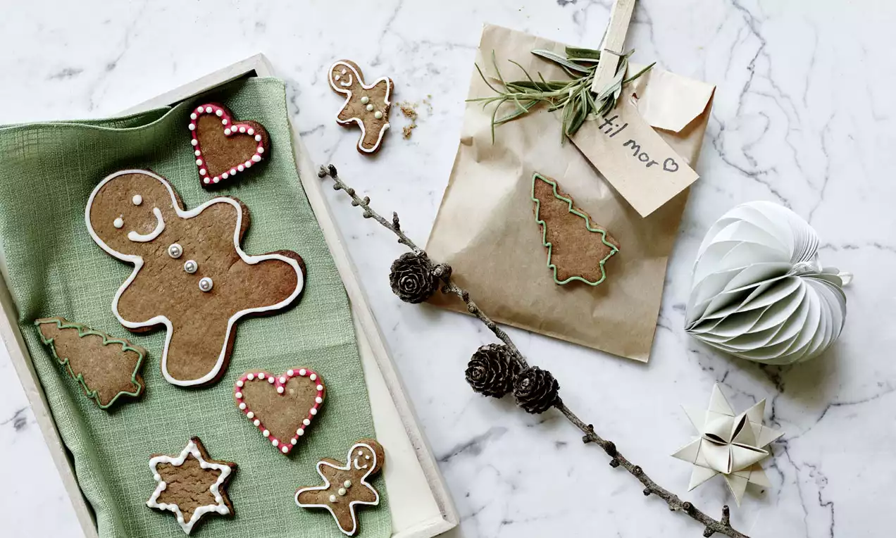 Klassiske brune julesmåkager formet som figurer med farvet glasur ligger på grønt klæde tæt på gavepose, grankogler og papirdekorationer