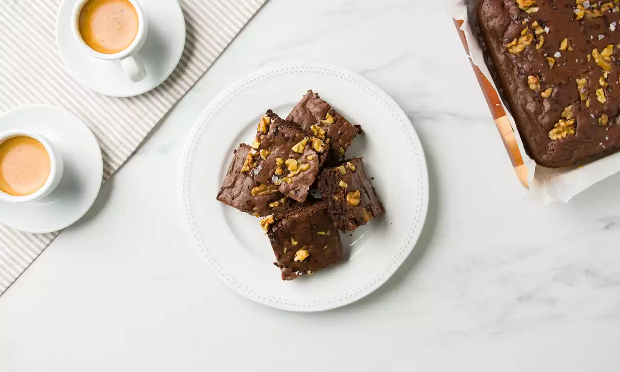 Cuadrados de brownie marrón oscuro con superficie ligeramente crujiente, trozos de nuez dorada encima y servidos en un plato blanco