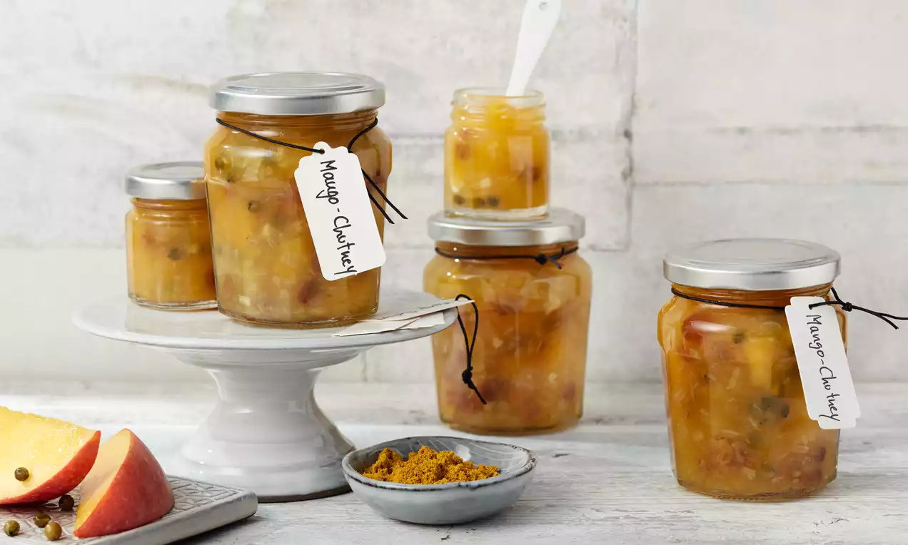 Goldgelbes Mango-Chutney mit stückiger Textur in Gläsern mit silbernem Deckel und handgeschriebenen Etiketten auf hellem Untergrund drapiert