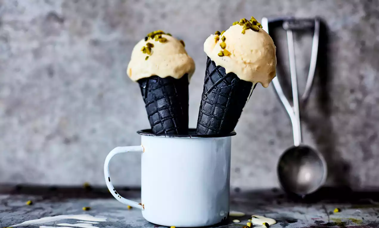 Înghețată de casă cu vanilie crem deschis pe cornete negre, presărată cu fistic mărunțit, servită într-o cană albă metalică