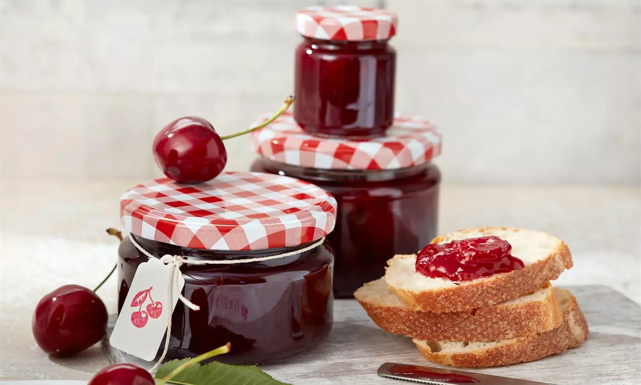 Kirschmarmelade im Glas mit tiefrot glänzender Oberfläche, auf Holzbrett, umgeben von frischen Kirschen und einem grünen Blatt