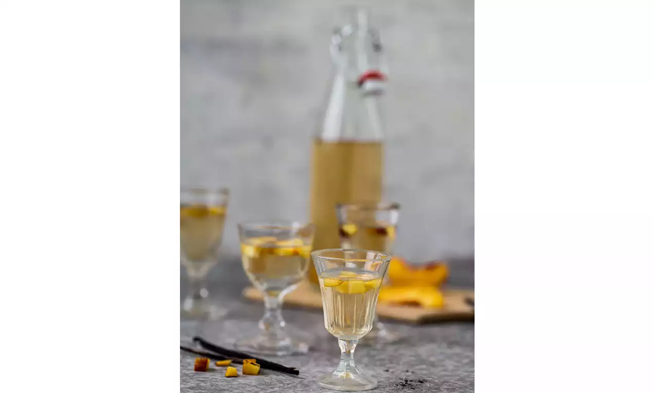 Klarer Aprikosen-Pfirsich-Likör in kleinen Gläsern mit gelben Fruchtwürfeln, dazu Flasche und frische Fruchtstücke auf grauem Untergrund