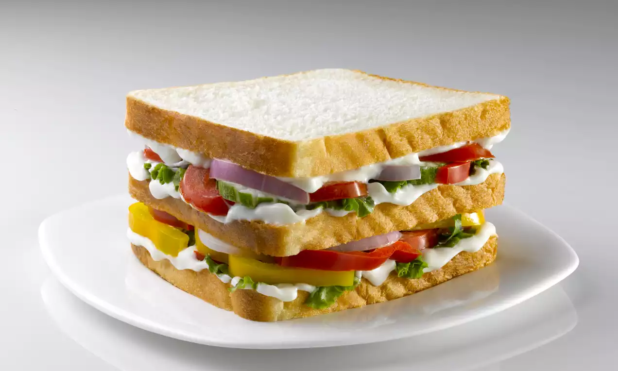 A triple-layer sandwich with golden toasted bread, crisp lettuce, tomato slices, onion rings, bell pepper and white cream on a white plate