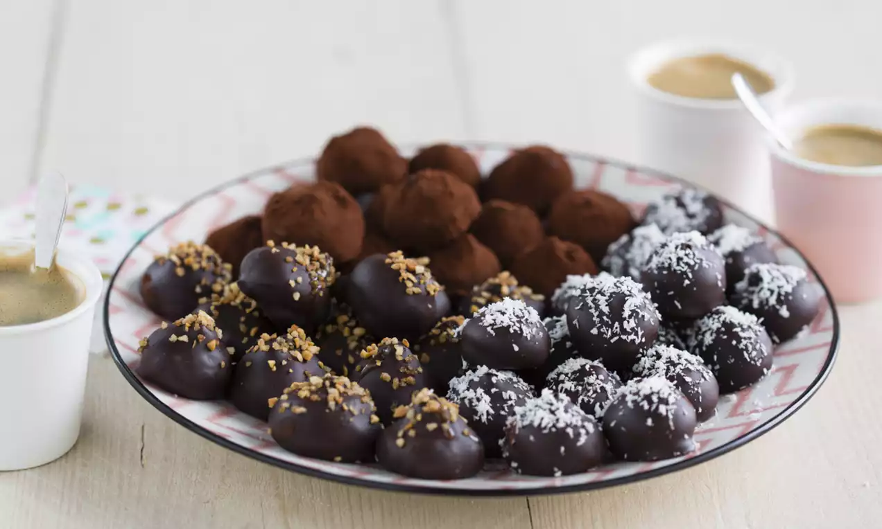 Ovale chocoladetruffels met cacao, nootjes en kokos, gerangschikt op een roze bord met drie kopjes koffie op een lichte houten tafel