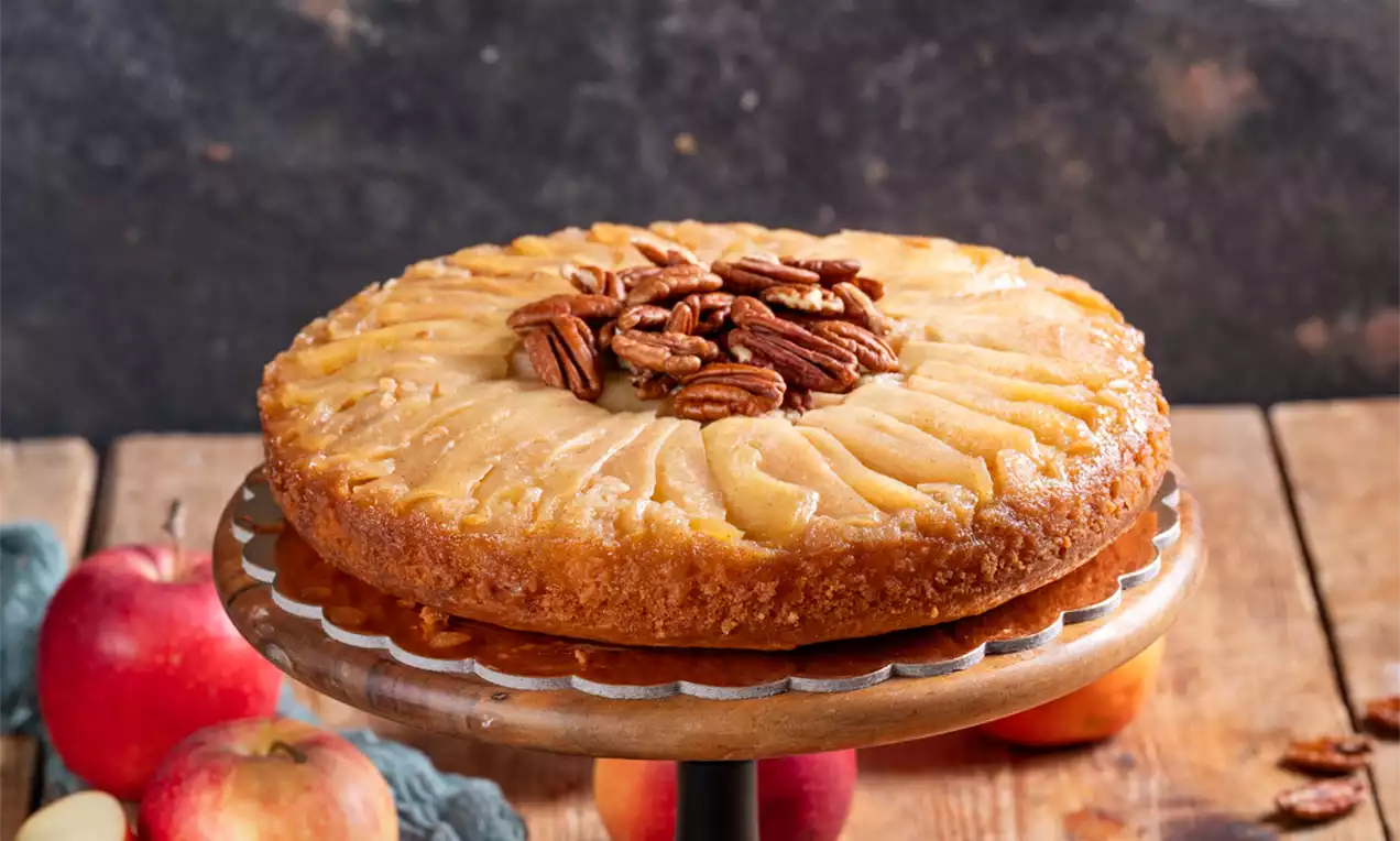 Zvrnjena jabolčna torta z zlato rjavo skorjo, urejenimi tankimi rezinami jabolk in pečenimi pekan orehi na kovinskem podstavku