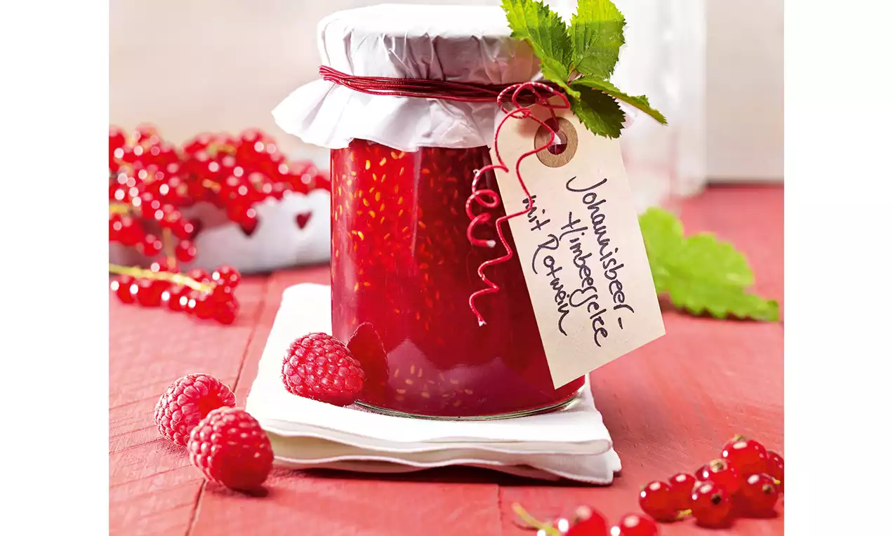 Glas mit leuchtend rotem Gelee, mit weißem Deckel und roter Kordel verziert, auf weißen Servietten neben Johannisbeeren und Himbeeren