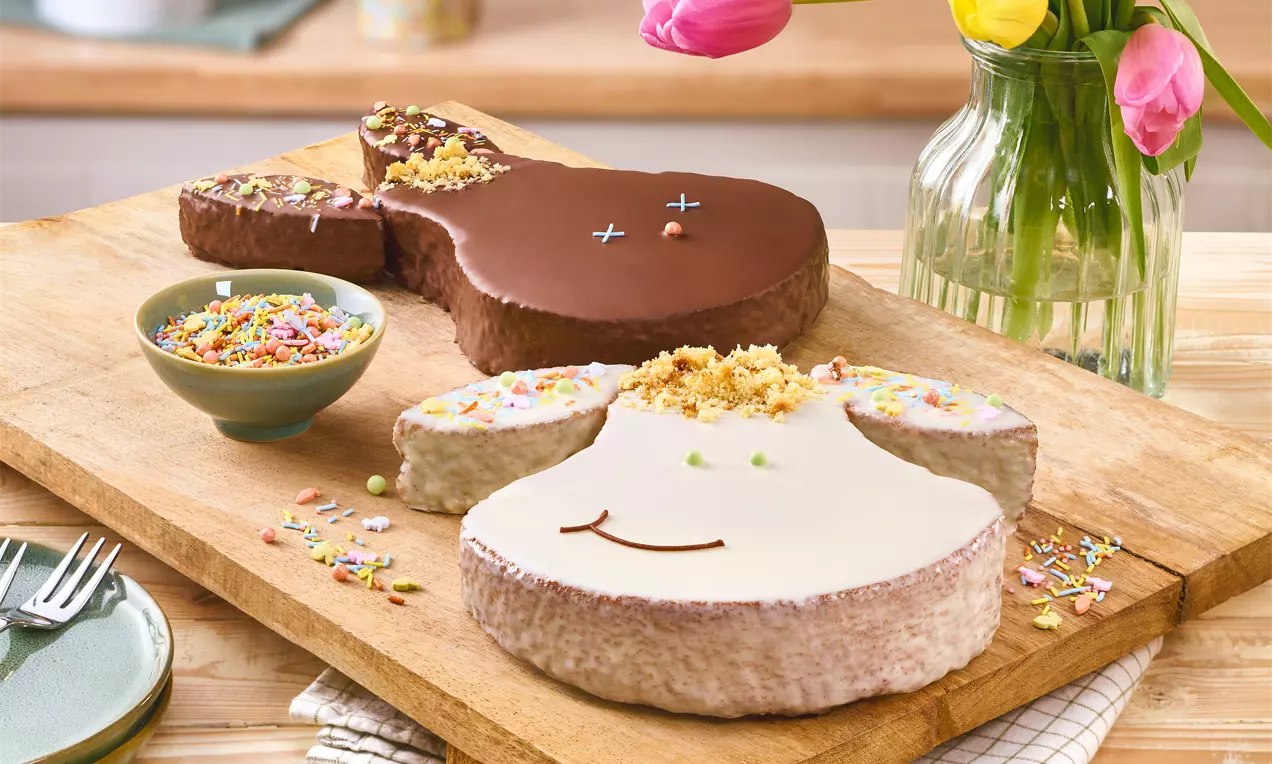 Zwei osterhasenförmige Kuchen mit Schokoglasur und heller Zuckerglasur, dekoriert mit Streuseln und Krümeln auf einem Holzbrett