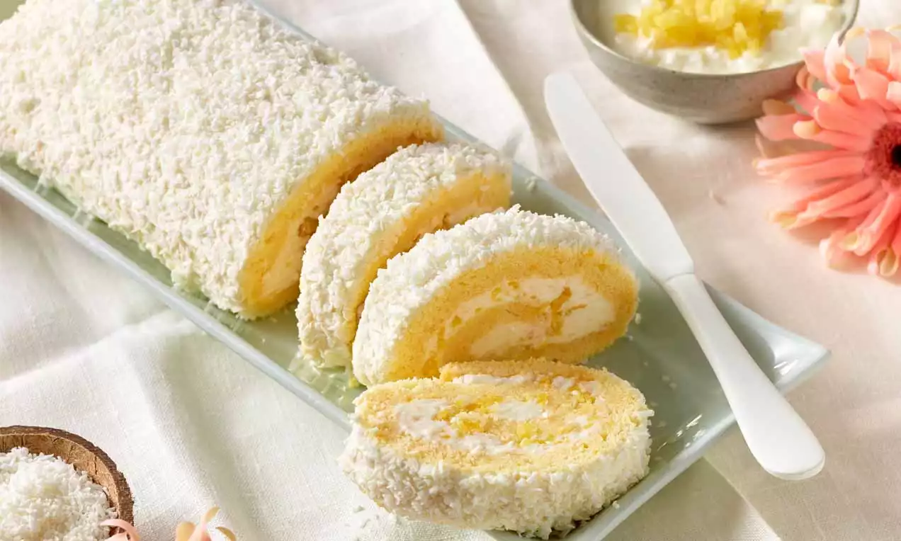 Gebrachte Ananas-Roulade mit gelber Füllung und weißem Kokosmantel, in Scheiben auf einer rechteckigen Platte mit Messer angerichtet